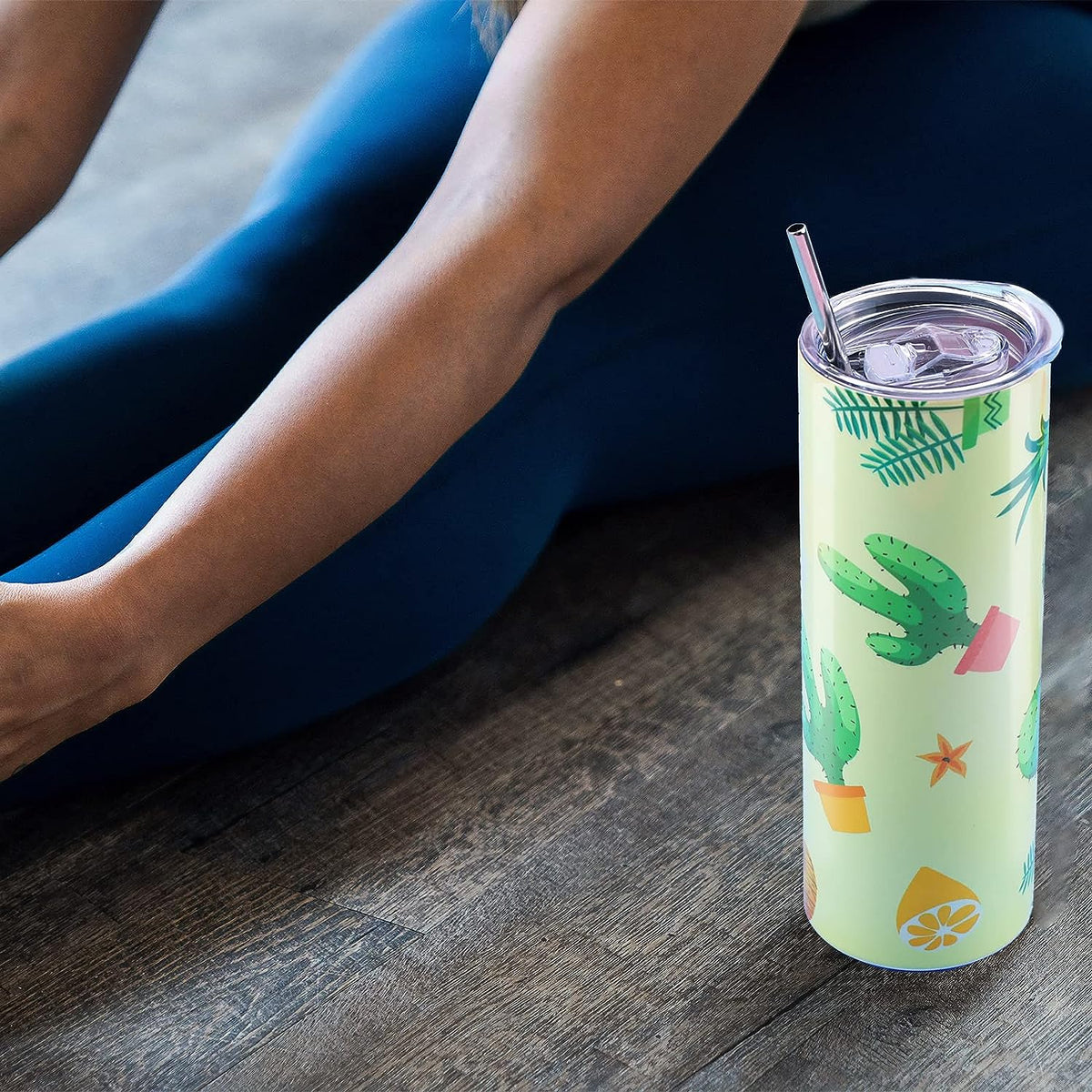Tumbler with cactus design on a wooden surface next to a person wearing blue leggings.